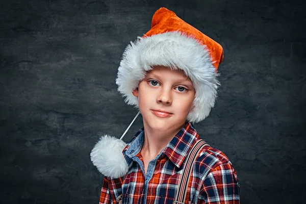 A handsome teen boy in Santas hat wearing a checkered shirt with suspenders. Isolated on a dark textured background. — Stock Photo, Image