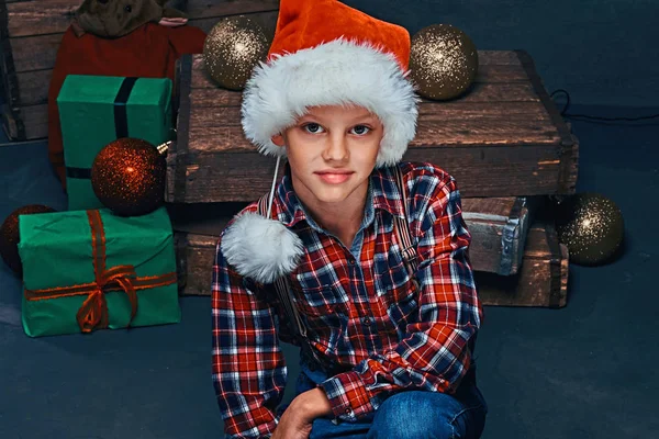 Adolescente em chapéu de Papai Noel vestindo uma camisa quadriculada sentada em uma sala com presentes de Natal, bolas e caixas de madeira . — Fotografia de Stock