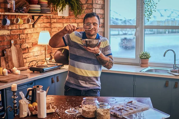Smiling man with a tattoo on his hand holding a bowl and inhaling the aroma of food in the kitchen.