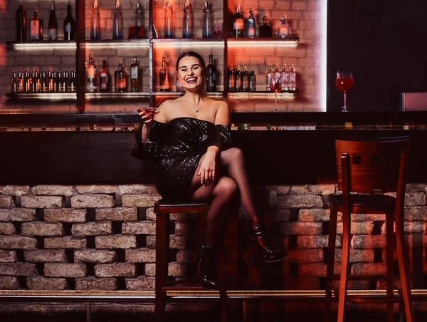 Uma mulher bonita alegre vestindo um vestido preto brilhante sentado em um balcão de bar — Fotografia de Stock