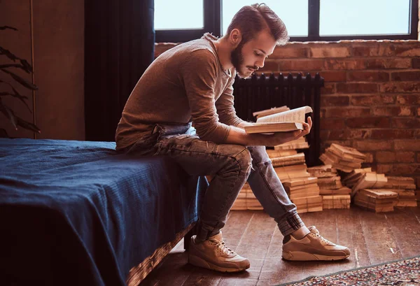 Student met stijlvolle baard en haren is voorbereiden op examens, zittend op het bed en het lezen van een boek. — Stockfoto