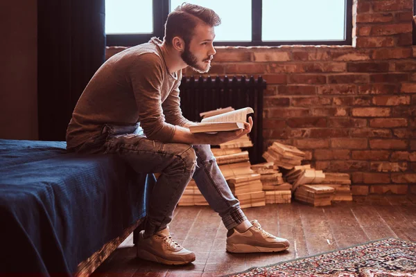 Estudante com barba elegante e cabelo está se preparando para exames, sentado na cama e lendo um livro . — Fotografia de Stock