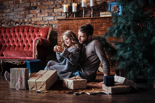 Happiness cute little girl holding christmas ball while sitting with her father on a floor surrounded by gifts — Stock Photo, Image