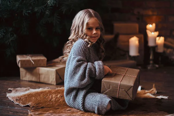 Cute little girl with blonde curly hair wearing a warm sweater holding a gift box while sitting on a floor next to the christmas tree. — Stock Photo, Image