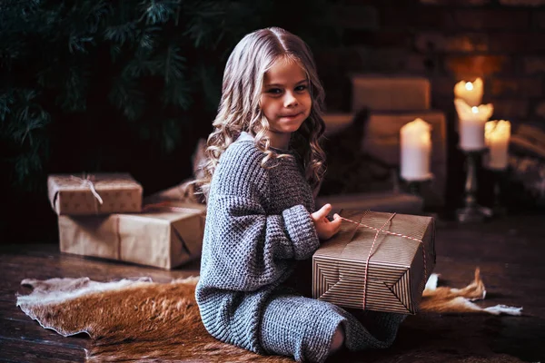 Linda niña con el pelo rubio rizado usando un suéter caliente sosteniendo una caja de regalo mientras está sentado en un piso al lado del árbol de Navidad . —  Fotos de Stock