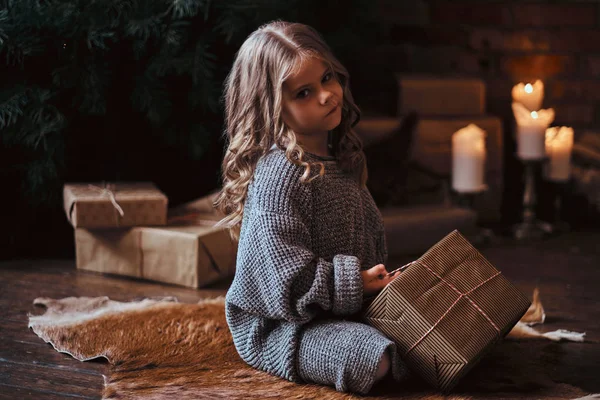 Dissatisfied little girl with blonde curly hair wearing a warm sweater sitting on a floor surrounded by gifts next to the christmas tree. — Stock Photo, Image