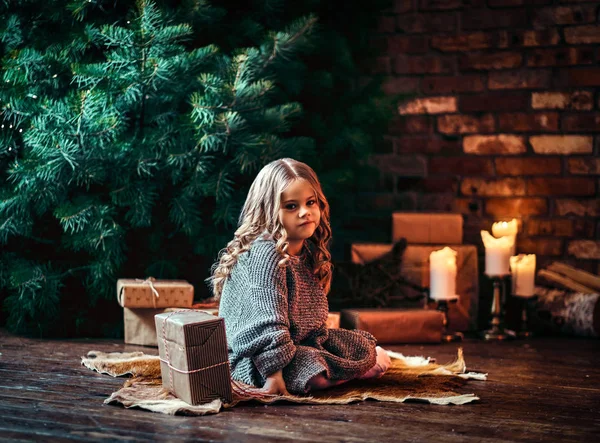 Dissatisfied little girl with blonde curly hair wearing a warm sweater sitting on a floor surrounded by gifts next to the christmas tree. — Stock Photo, Image