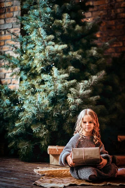 Cute little girl with blonde curly hair wearing a warm sweater holding a gift box while sitting on a floor next to the christmas tree. — Stock Photo, Image