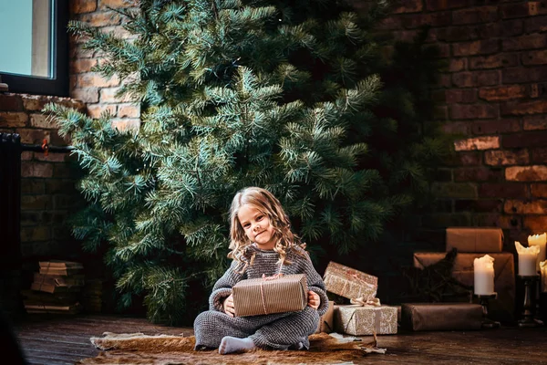Счастливая маленькая девочка с кудрявыми светлыми волосами в тёплом свитере с подарочной коробкой на полу рядом с рождественской ёлкой . — стоковое фото