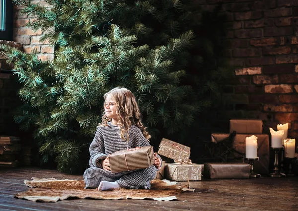 ギフト用の箱を押しながらクリスマス ツリーの横にある床の上に座って暖かいセーターを着ている金髪の巻き毛を持つ幸せな少女. — ストック写真