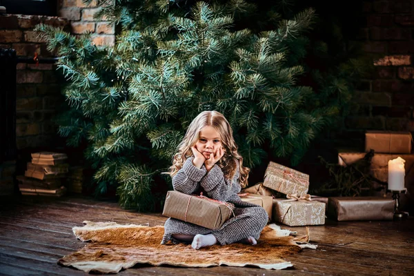 Dissatisfied little girl with blonde curly hair wearing a warm sweater sitting on a floor surrounded by gifts next to the christmas tree. — Stock Photo, Image