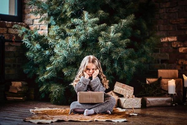 Cute little girl with blonde curly hair wearing a warm sweater holding a gift box while sitting on a floor next to the christmas tree. — Stock Photo, Image
