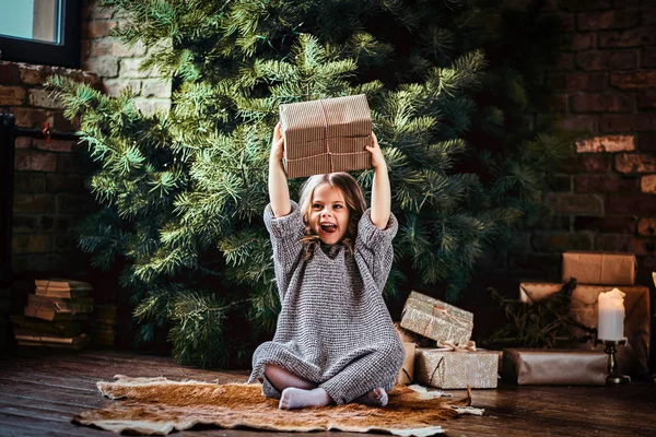 Joyful little girl with blonde curly hair wearing a warm sweater throws up a gift boxes while sitting on a floor next to the christmas tree. — Stock Photo, Image