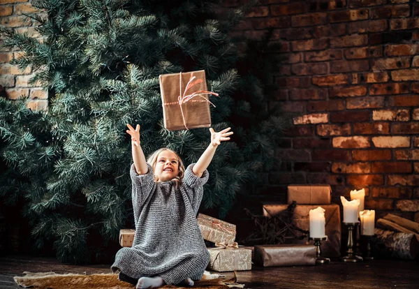 暖かいセーターを着ている金髪の巻き毛のうれしそうな小さな女の子がクリスマス ツリーの横にある床の上に座っている間ギフト ボックスをスローします。. — ストック写真