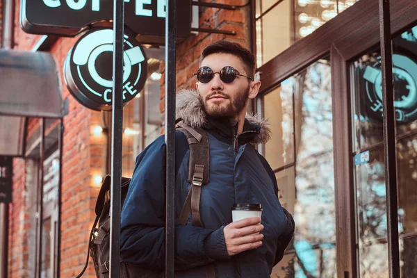 Un joven guapo con una mochila sosteniendo una taza de café mientras estaba parado cerca de un café afuera . —  Fotos de Stock