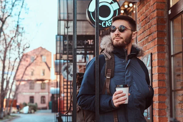 Een knappe jongeman met een rugzak die houden van een kopje koffie terwijl je in de buurt van een café buiten. — Stockfoto