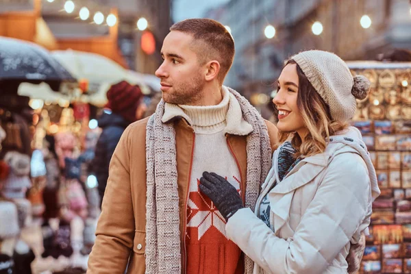 Um jovem casal romântico andando na rua na época do Natal, de pé perto de lojas decoradas à noite, desfrutando de passar o tempo juntos . — Fotografia de Stock