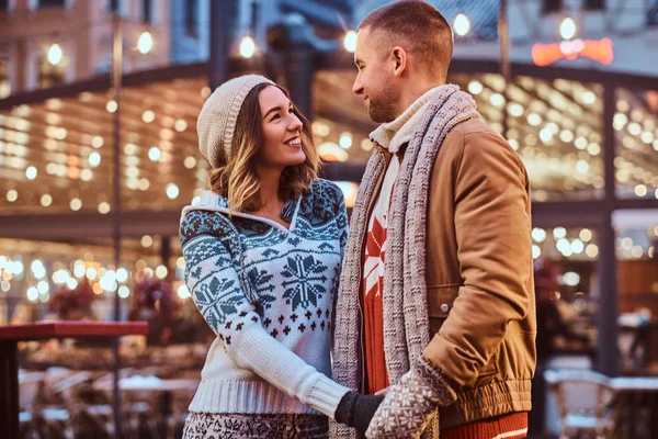 A young romantic couple holding hands and looking at each other while standing on the street at Christmas time, enjoying spending time together. — Stock Photo, Image
