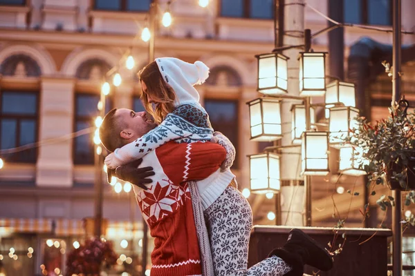 Young romantic couple enjoying spending time together at Christmas on street decorated with beautiful lights. A happy young guy raised his girlfriend. — Stock Photo, Image