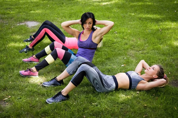 Tres amigas deportivas hacen ejercicio en el parque al aire libre, tumbadas en la hierba y haciendo ejercicios de abdominales —  Fotos de Stock