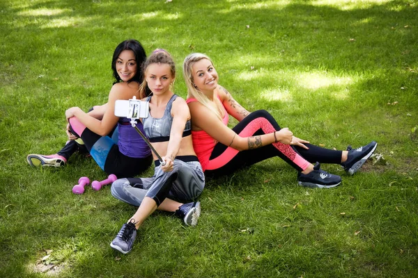 Tre amiche sportive sedute su un'erba al parco all'aperto, fanno selfie al telefono con un bastone — Foto Stock