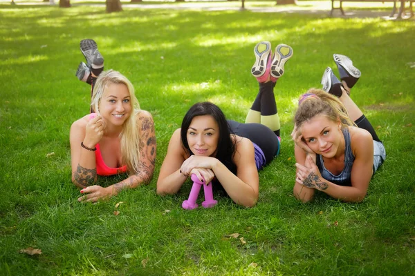 Three sportive female friends lie on the grass at the park outdoors, relaxing after workout, looking at camera. — Stock Photo, Image