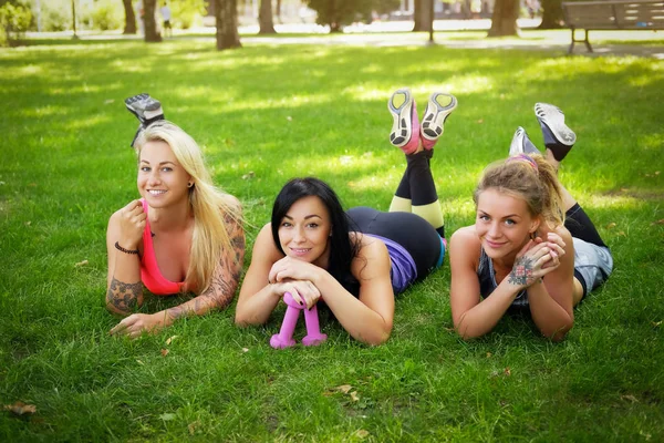 Drie sportieve vriendinnen liggen op het gras in het park buiten, ontspannen na training, camera kijken. — Stockfoto