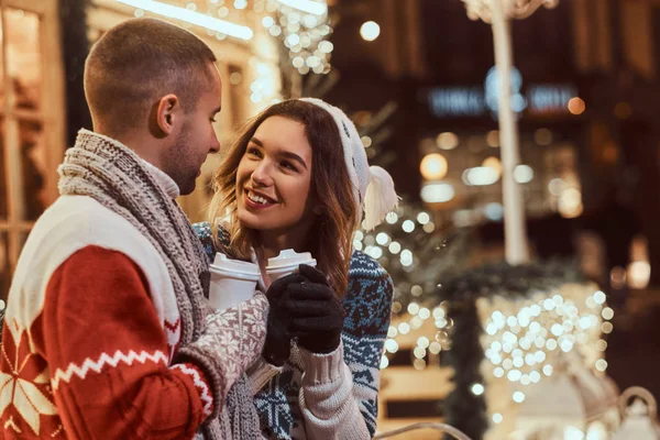 Um jovem casal romântico vestindo roupas quentes ao ar livre na época do Natal, sentado em um banco e aquecendo com café quente na rua da noite decorado com belas luzes . — Fotografia de Stock