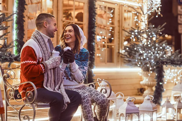 Una joven pareja romántica que viste ropa de abrigo al aire libre en Navidad, sentada en un banco y calentándose con café caliente en la calle de la noche decorada con hermosas luces . — Foto de Stock