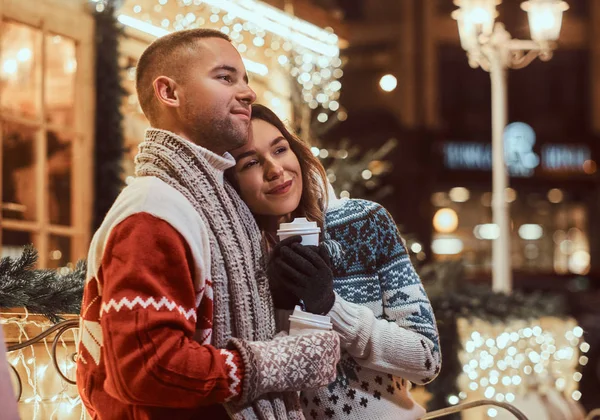 Una joven pareja romántica que viste ropa de abrigo al aire libre en Navidad, sentada en un banco y calentándose con café caliente en la calle de la noche decorada con hermosas luces . — Foto de Stock