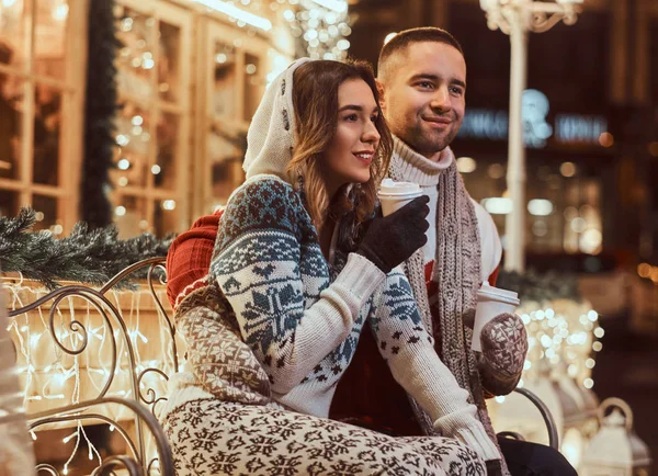 젊은 낭만적인 커플 크리스마스 시간에 야외 따뜻한 옷을 입고, 아름 다운 조명으로 장식 하는 벤치에 앉아서 저녁 거리에 뜨거운 커피와 온난화. — 스톡 사진