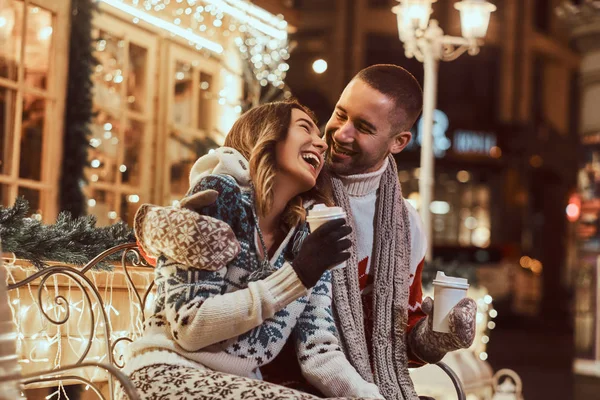 Um jovem casal romântico vestindo roupas quentes ao ar livre na época do Natal, sentado em um banco e aquecendo com café quente na rua da noite decorado com belas luzes . — Fotografia de Stock
