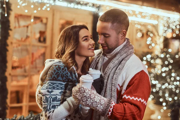 Um jovem casal romântico vestindo roupas quentes ao ar livre na época do Natal, abraçando e aquecendo com café quente na rua da noite decorada com belas luzes . — Fotografia de Stock