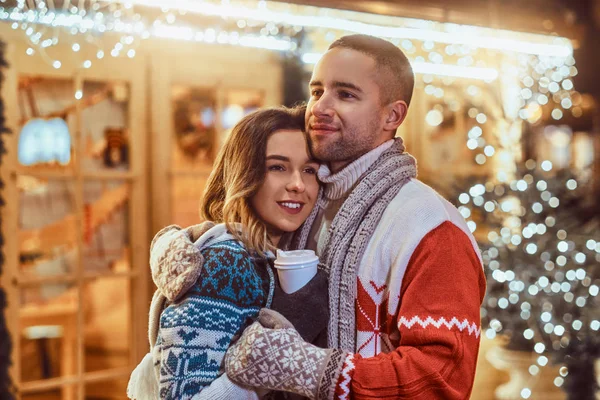 Una joven pareja romántica que viste ropa de abrigo al aire libre en Navidad, abrazándose y calentándose con café caliente en la calle de la noche decorada con hermosas luces . — Foto de Stock