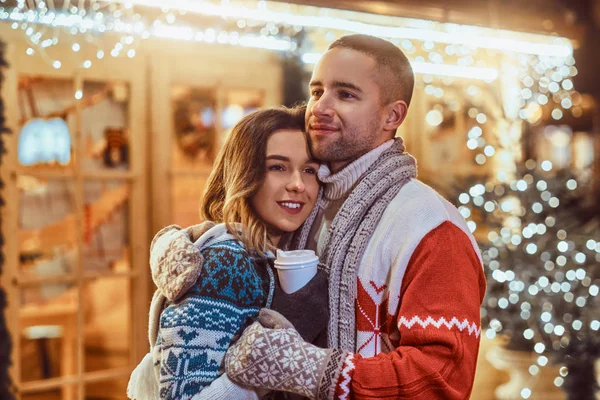 Um jovem casal romântico vestindo roupas quentes ao ar livre na época do Natal, abraçando e aquecendo com café quente na rua da noite decorada com belas luzes . — Fotografia de Stock