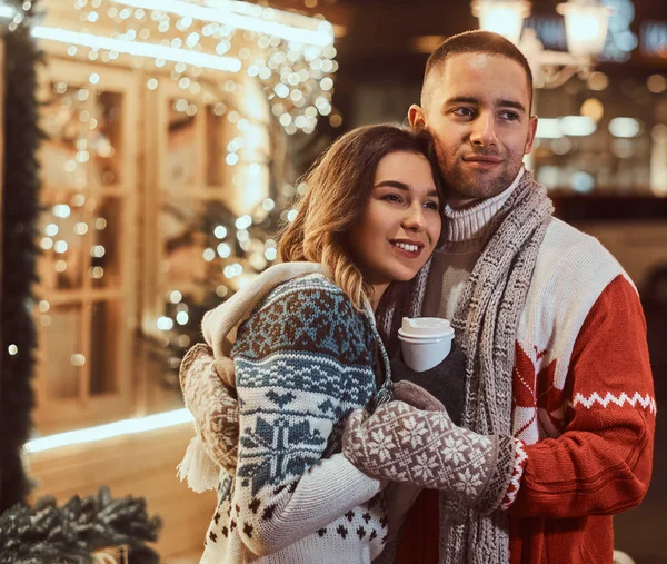 Una joven pareja romántica que viste ropa de abrigo al aire libre en Navidad, abrazándose y calentándose con café caliente en la calle de la noche decorada con hermosas luces . — Foto de Stock