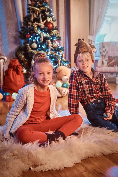Счастливые брат и сестра сидят на меховом ковре возле рождественской елки дома . — стоковое фото