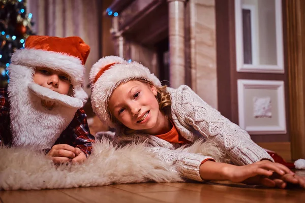 幸福的兄妹戴着圣诞老人的帽子躺在家里一棵圣诞树附近的皮草地毯上. — 图库照片