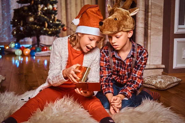 Happy brother and sister wearing Santa and deer hats sitting on a fur carpet and opens gift near a Christmas tree at home.