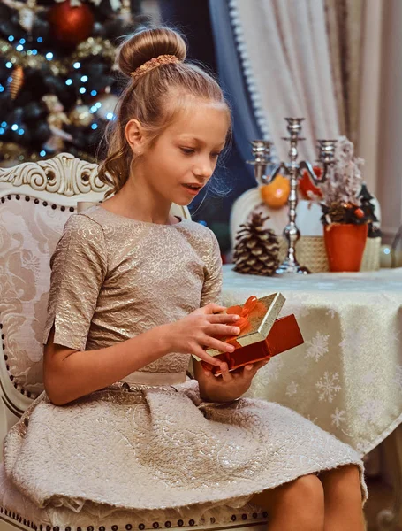 Menina ute vestindo um vestido bonito sentado em uma cadeira e abre uma caixa de presente — Fotografia de Stock