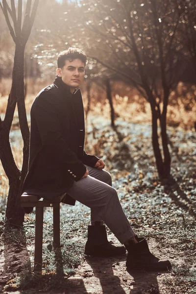 Un joven vistiendo un abrigo negro sentado en un banco en el bosque de otoño — Foto de Stock