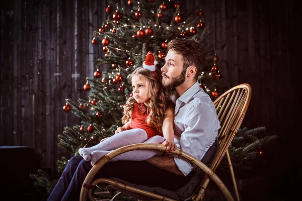 Dad and daughter sitting together on a rocking chair near a Christmas tree at home.