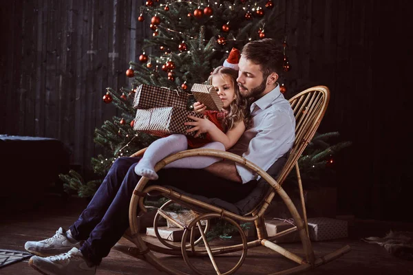 Baba ve kızı birlikte bir sallanan sandalyede otururken hediye kutuları tutarak bir Noel ağacı evde yakın:. — Stok fotoğraf