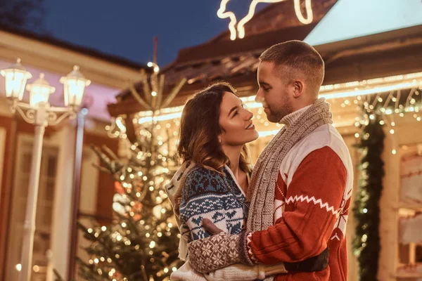 Una joven pareja romántica con ropa abrigada abrazándose al aire libre en Navidad, de pie en la calle de la noche decorada con hermosas luces . —  Fotos de Stock