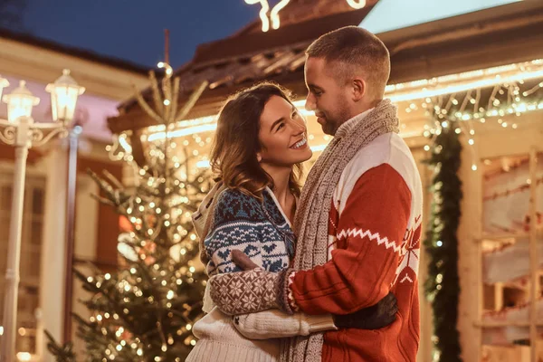 Um jovem casal romântico vestindo roupas quentes abraçando ao ar livre na época do Natal, de pé na rua da noite decorada com belas luzes . — Fotografia de Stock