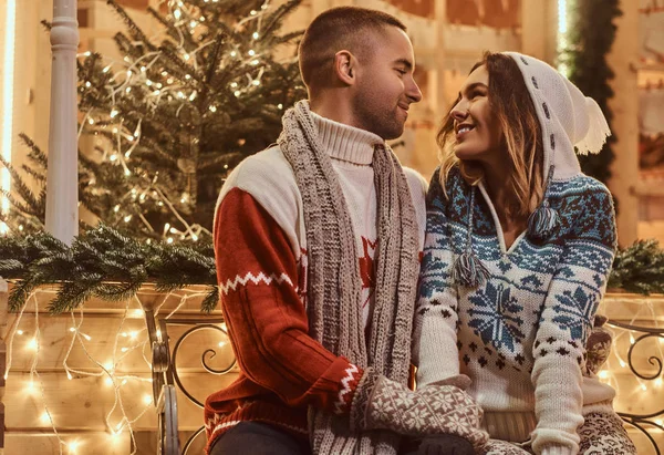 Um jovem casal romântico vestindo roupas quentes ao ar livre na época do Natal, sentado em um banco na rua à noite decorado com belas luzes . — Fotografia de Stock