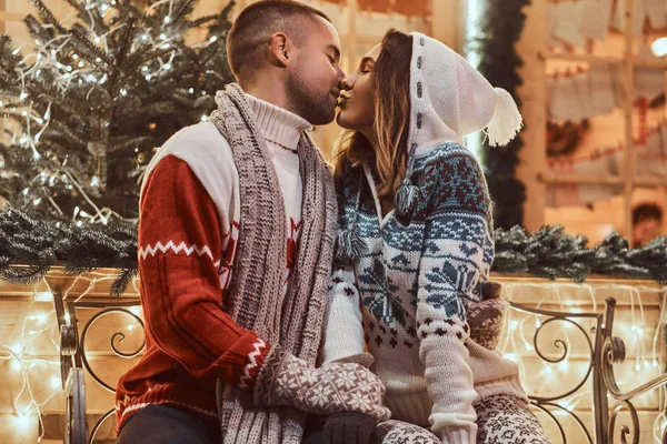 Una joven pareja romántica que viste ropa de abrigo al aire libre en Navidad, sentada en un banco en la calle de la noche decorada con hermosas luces . —  Fotos de Stock