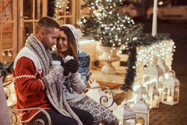 Una joven pareja romántica que viste ropa de abrigo al aire libre en Navidad, sentada en un banco y calentándose con café caliente en la calle de la noche decorada con hermosas luces . — Foto de Stock