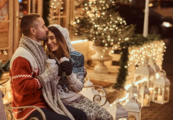 Um jovem casal romântico vestindo roupas quentes ao ar livre na época do Natal, sentado em um banco e aquecendo com café quente na rua da noite decorado com belas luzes . — Fotografia de Stock