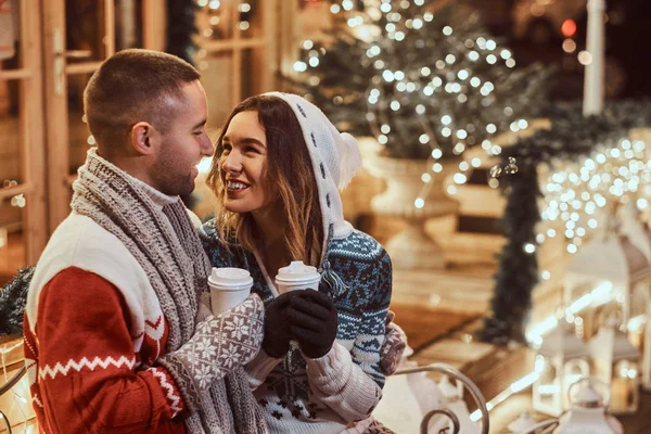 Una joven pareja romántica que viste ropa de abrigo al aire libre en Navidad, sentada en un banco y calentándose con café caliente en la calle de la noche decorada con hermosas luces . — Foto de Stock